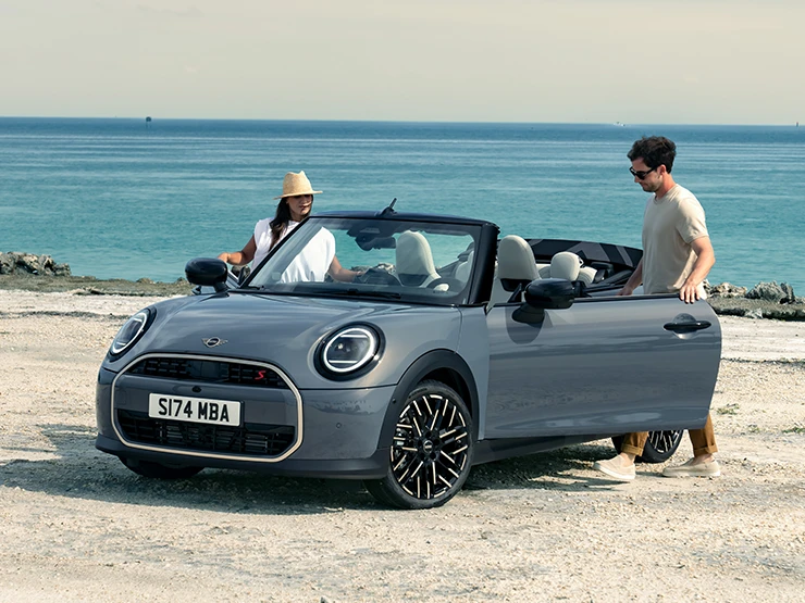 Eestvaade vasakult: rannas seisev MINI Cooper Convertible värvitoonis Copper Grey, auto kõrval on kaks inimest.