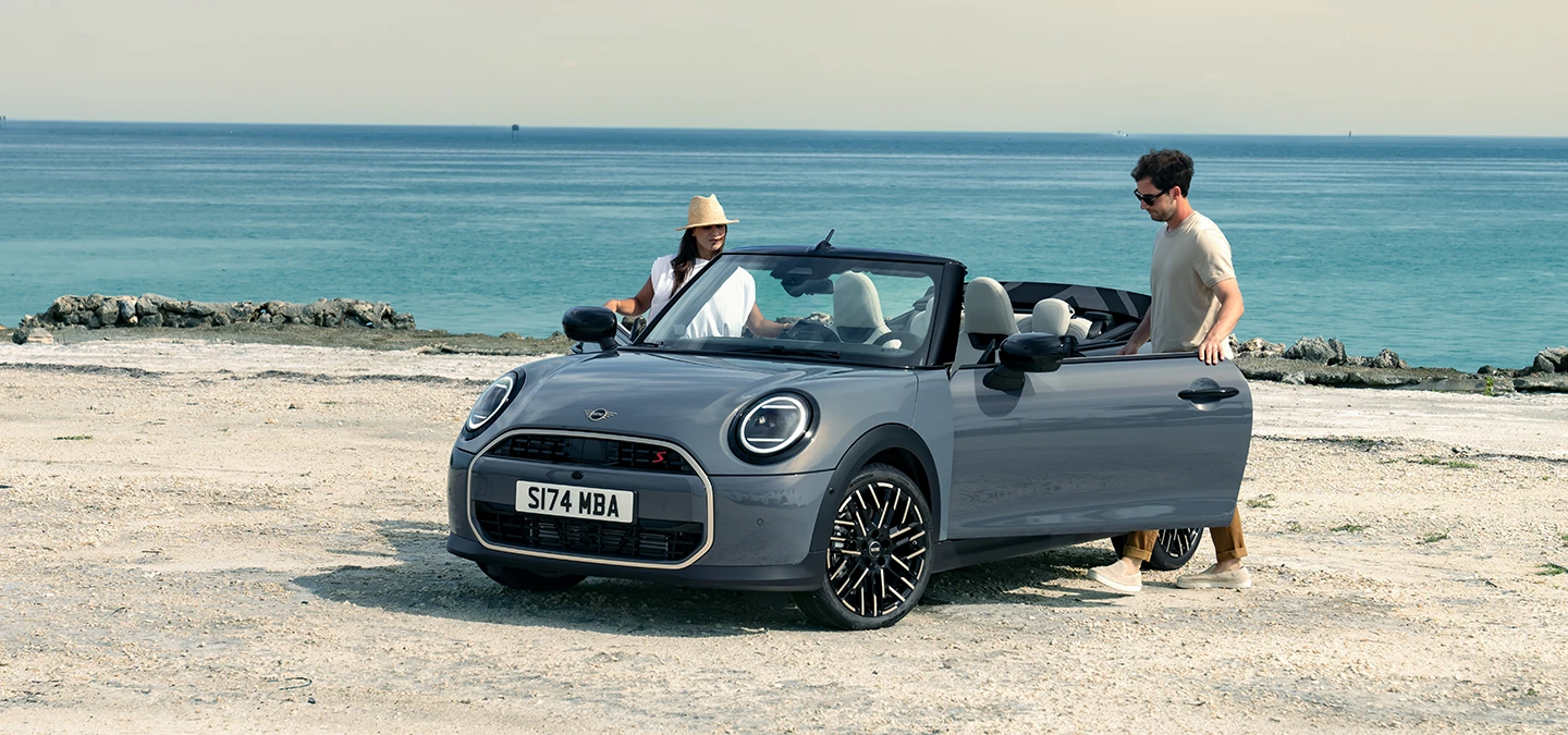 Eestvaade vasakult: rannas seisev MINI Cooper Convertible värvitoonis Copper Grey, auto kõrval on kaks inimest.