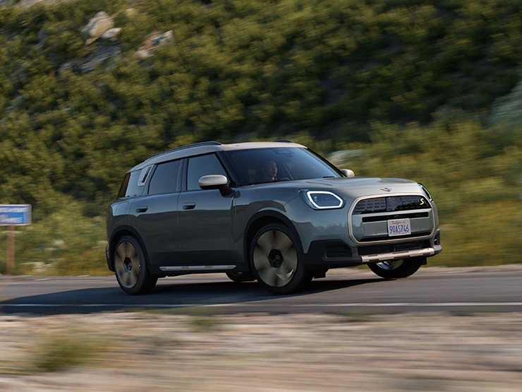 Smokey Greeni värvitoonis ja Vibrant Silveri katusega täiselektriline MINI Countryman maastikul sõitmas, külgvaade.