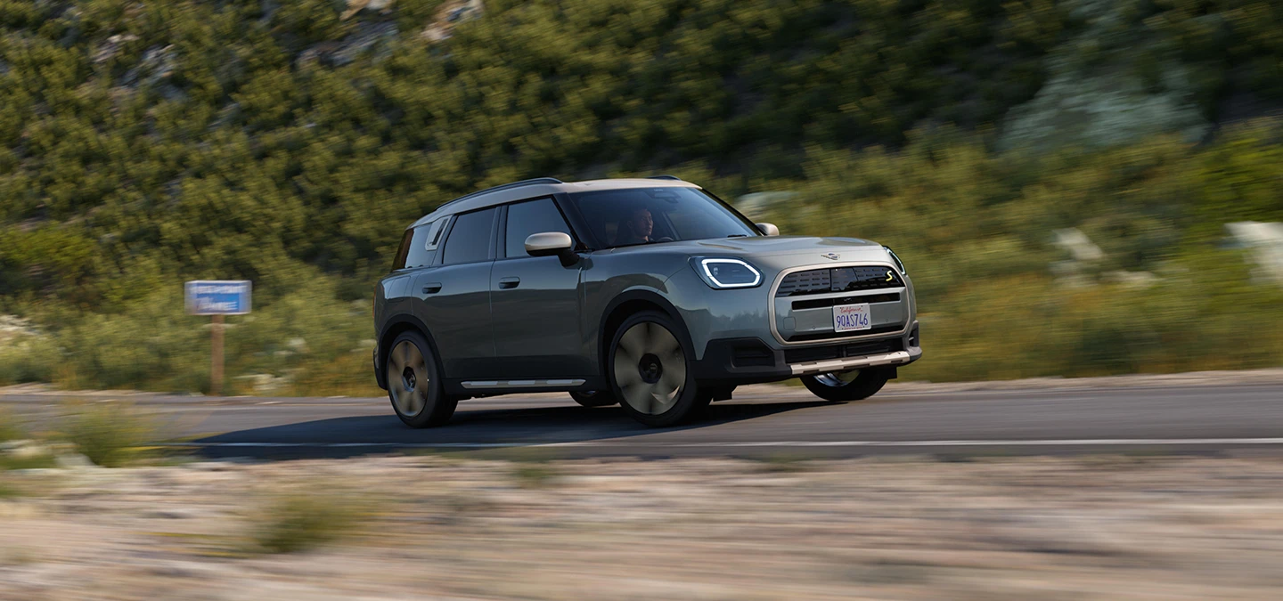 Smokey Greeni värvitoonis ja Vibrant Silveri katusega täiselektriline MINI Countryman maastikul sõitmas, külgvaade.