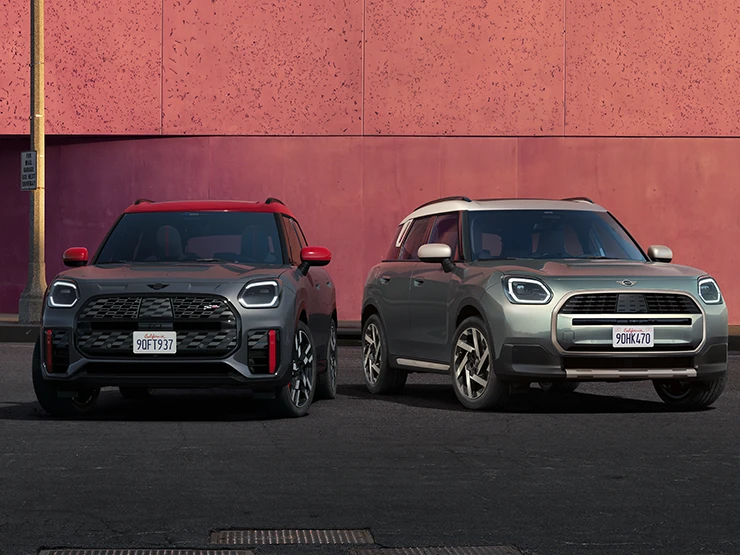 Eestvaade Legend Grey värvitoonis ja punase katusega MINI Countryman JCW-le ning Smokey Greeni värvitoonis ja Vibrant Silveri katusega MINI Countrymanile, mis seisavad kõrvuti.