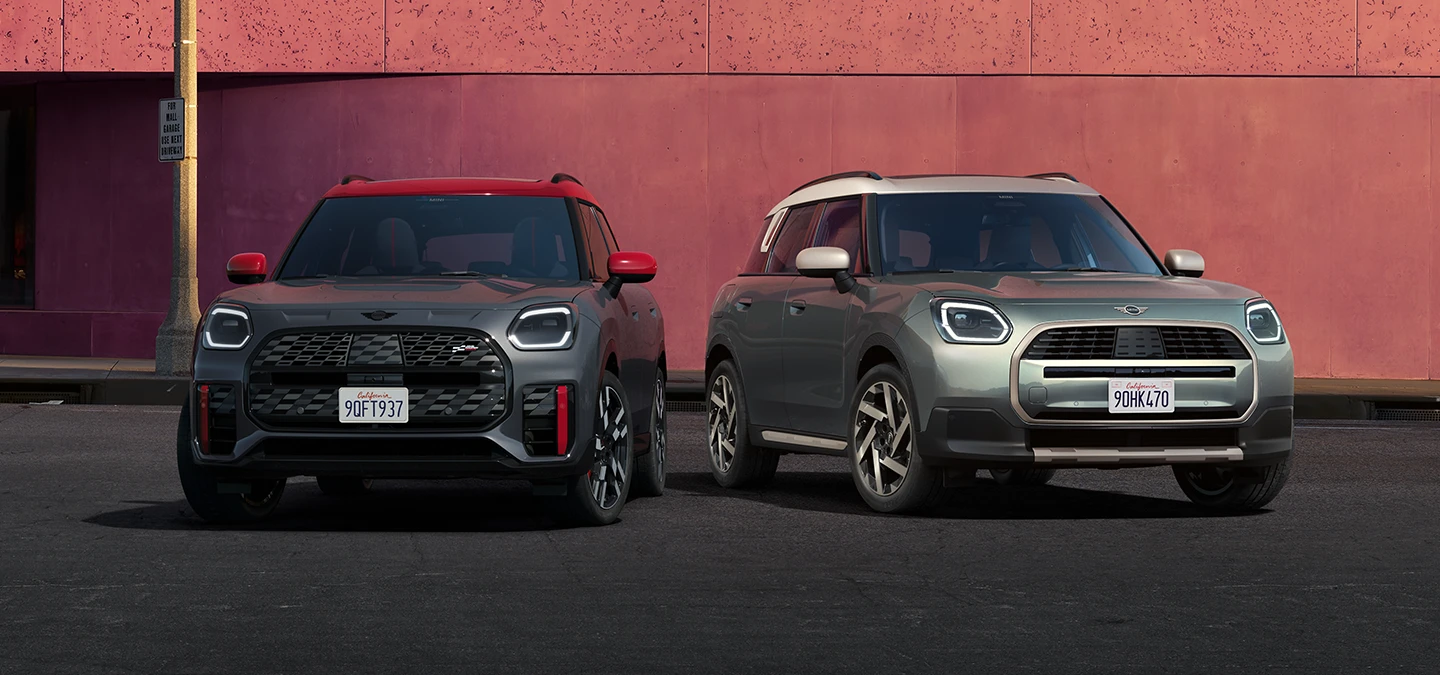 Eestvaade Legend Grey värvitoonis ja punase katusega MINI Countryman JCW-le ning Smokey Greeni värvitoonis ja Vibrant Silveri katusega MINI Countrymanile, mis seisavad kõrvuti.