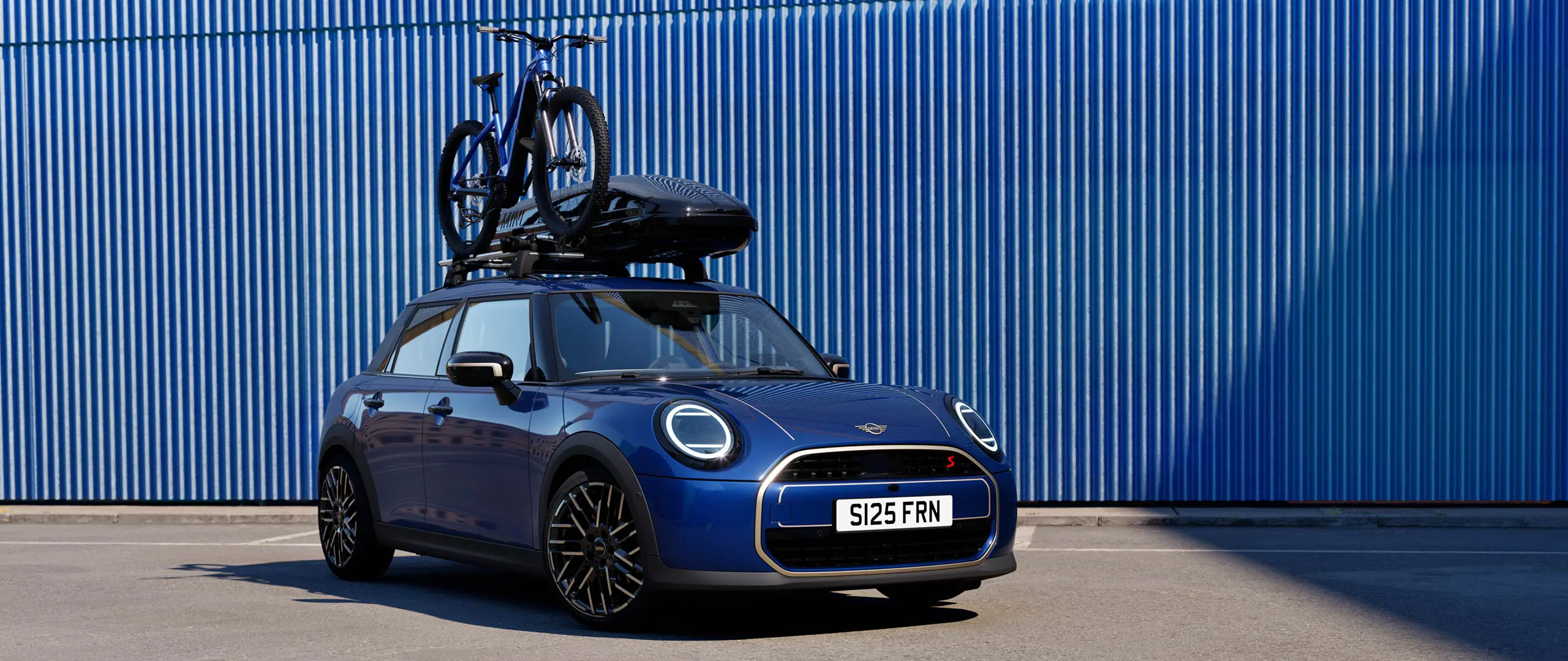 MINI Aceman esivaates värvitoonis Indigo Sunset Blue koos MINI katusele paigaldatava jalgrattahoidikuga, millel on maastikuratas, ja MINI katuseboksiga 320.